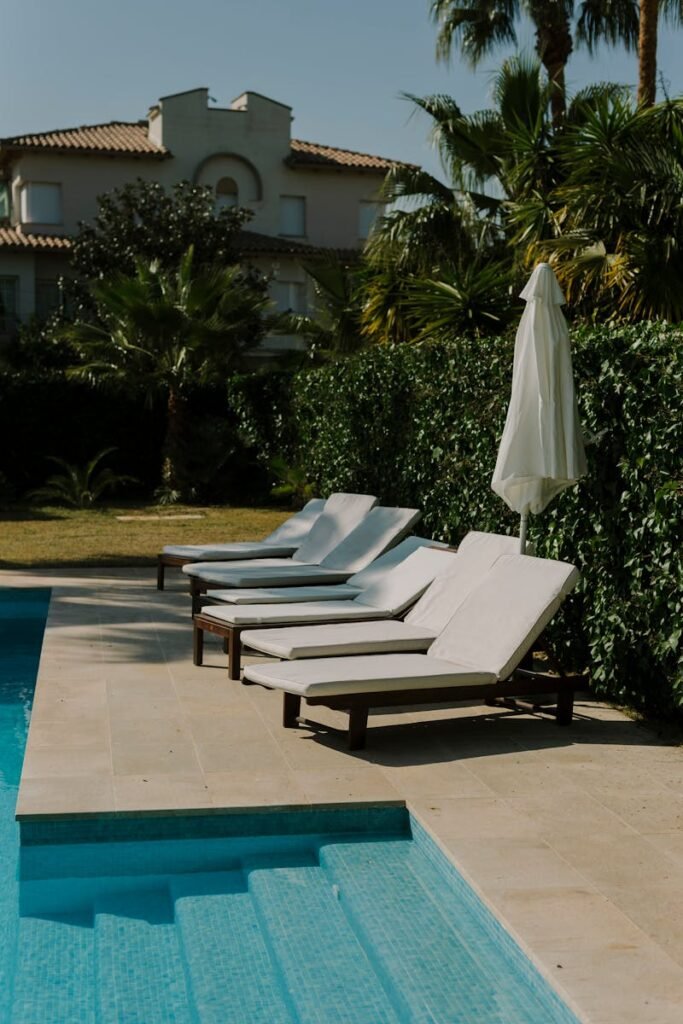 pexels-photo-15780455-15780455 Sunlit poolside with cushioned lounge chairs and lush greenery creates a perfect summer retreat.